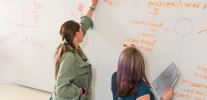两个学生用记号笔在白板上写字. 黑板上写着“正式评估”和“教练vs .教练”. 卡式肺囊虫肺炎."
