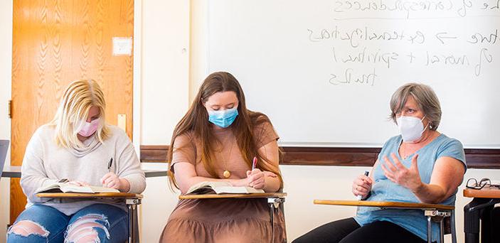 Dr. Ann Brunjes sits at a desk teaching in front of a white board that shows partial notes that read "correspondences" and " externalizations of moral and spiritual truths." Two students sit at desks beside her taking notes in their books.